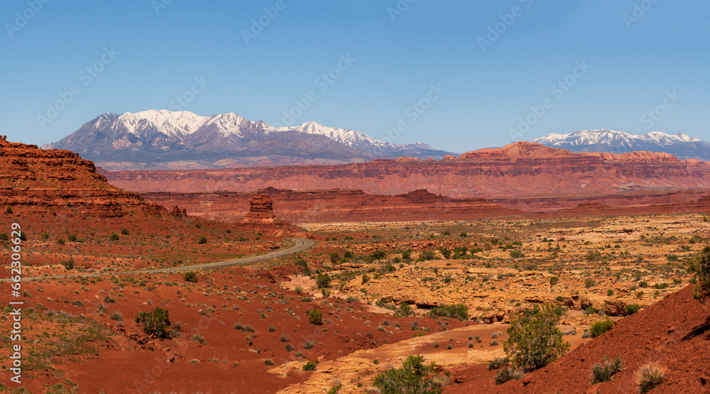 Panoramic view of a desert road in southern Utah, USA with snowy mountains in the horizon and clear blue sky.