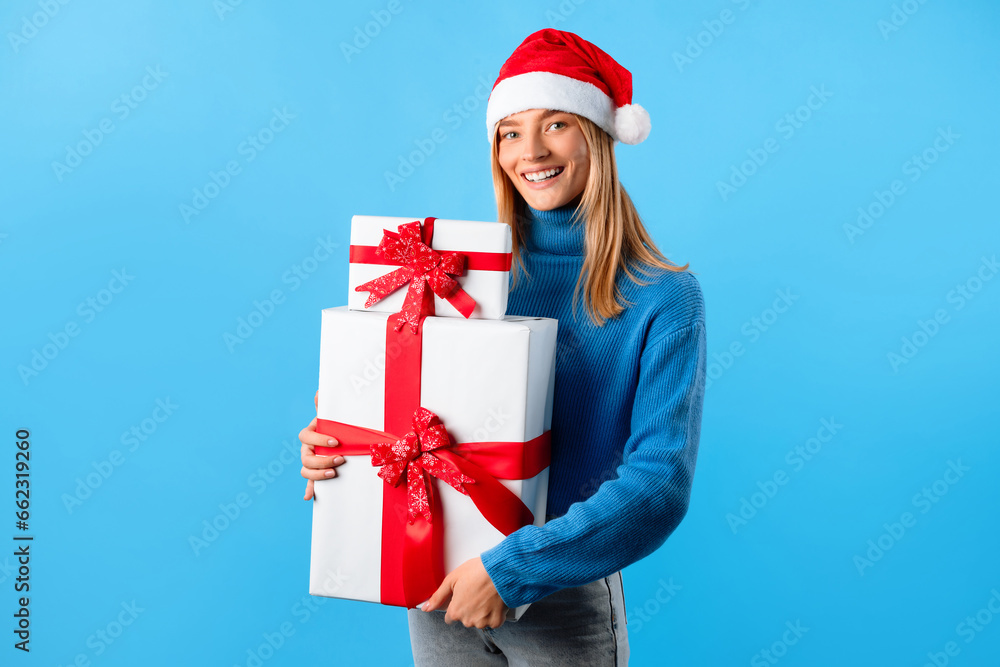 Glad woman with Santa hat holding Christmas gift boxes and smiling at camera, standing isolated on blue background Glad woman with Santa hat holding Christmas gift boxes and smiling at camera, standing isolated on blue background
