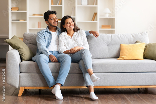 Happy dreamy indian couple looking aside at free space while sitting on sofa