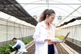 Happy smiling beautiful Asian botanist scientist woman in lab coat holding laptop computer, female biological researcher working on experimental plant plots in greenhouse. Biology Agricultural Science