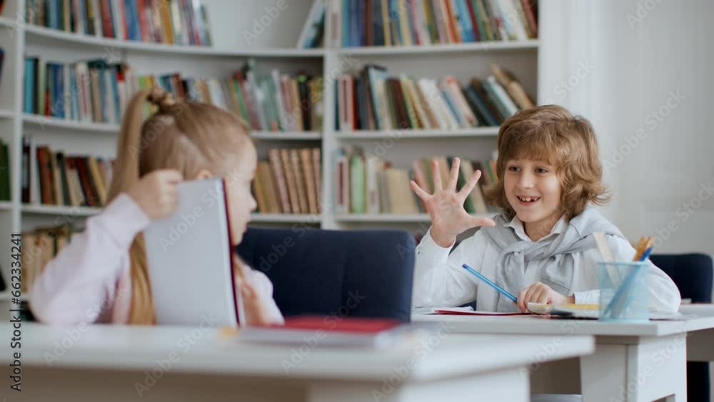 Pensive little girl having troubles with math test, turning to classmate and ask him about answer, boy gesturing five Pensive little girl having troubles with math test, turning to classmate and ask him about answer, boy gesturing five