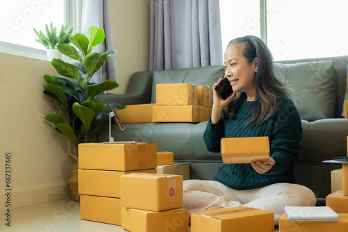 Senior Asian woman selling products online, small business owner starting an SME. Elderly woman using a smartphone or tablet taking and checking online orders to prepare boxes of products.