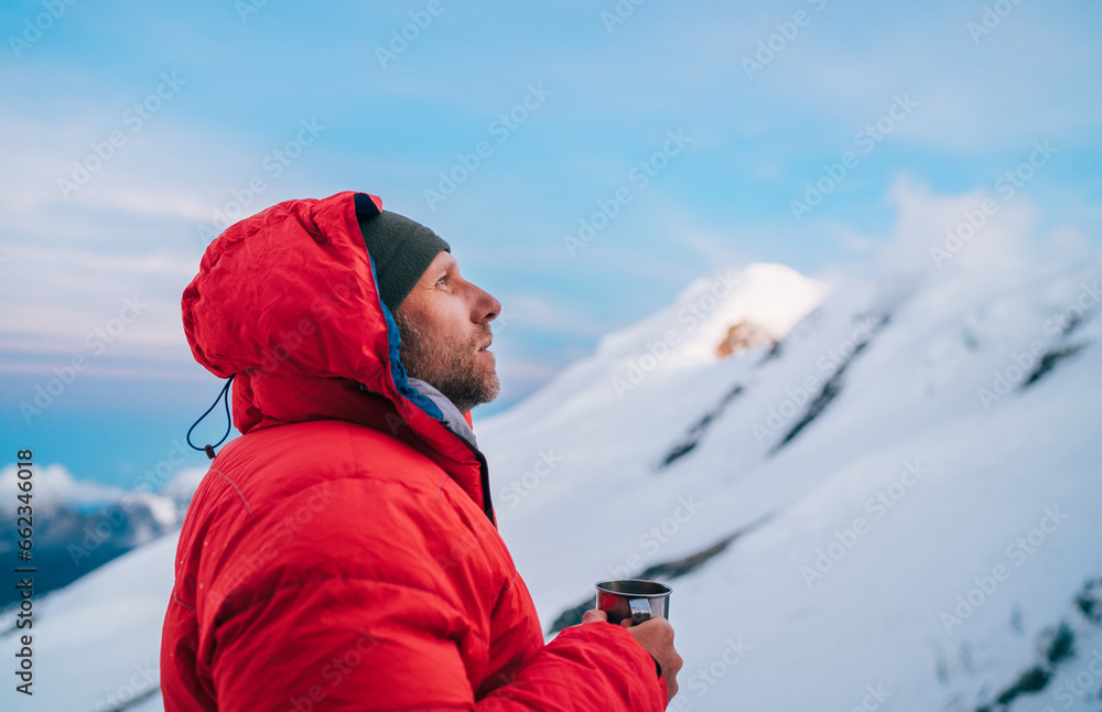 Portrait of high altitude mountaineer dressed red warm dawn jacket holding metal mug of hot tea thinking and enjoying fresh mountains frosty air. Mera Peak High Camp 5700m, Himalayas, Nepal.