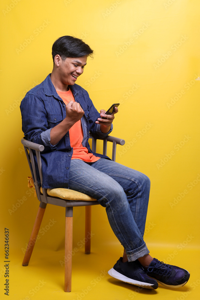 Young Asian man clenched fist showing victory gesture while using smartphone over yellow background