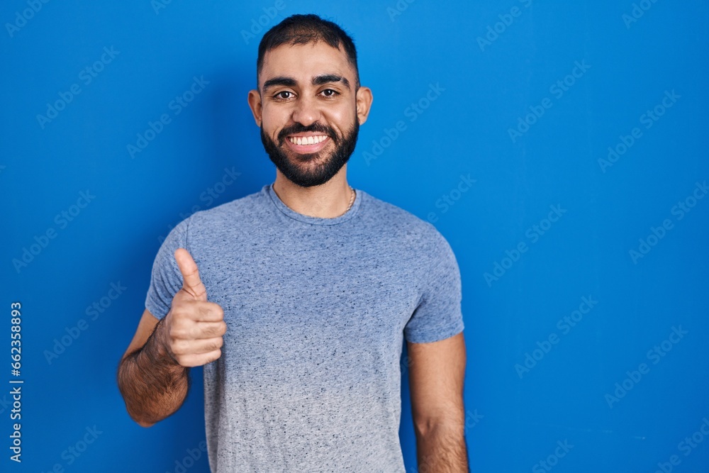 Middle east man with beard standing over blue background doing happy thumbs up gesture with hand. approving expression looking at the camera showing success. Middle east man with beard standing over blue background doing happy thumbs up gesture with hand. approving expression looking at the camera showing success.