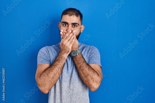 Middle east man with beard standing over blue background shocked covering mouth with hands for mistake. secret concept.