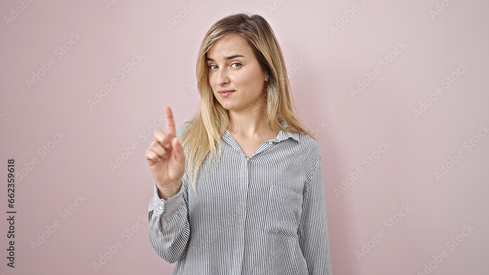 Young blonde woman standing with serious expression saying no with finger over isolated pink background