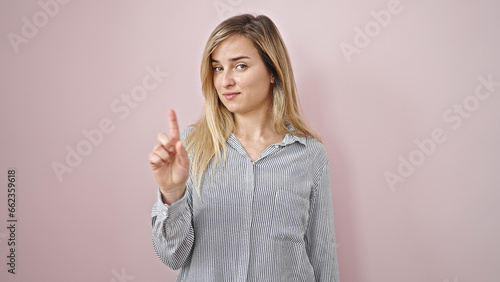 Young blonde woman standing with serious expression saying no with finger over isolated pink background
