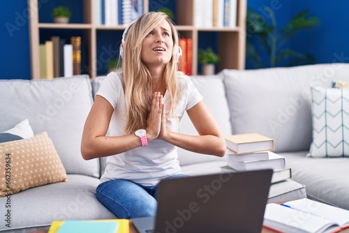 Young blonde woman studying using computer laptop at home begging and praying with hands together with hope expression on face very emotional and worried. begging.