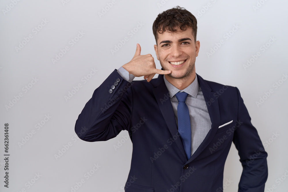 Young hispanic business man wearing suit and tie smiling doing phone gesture with hand and fingers like talking on the telephone. communicating concepts. Young hispanic business man wearing suit and tie smiling doing phone gesture with hand and fingers like talking on the telephone. communicating concepts.