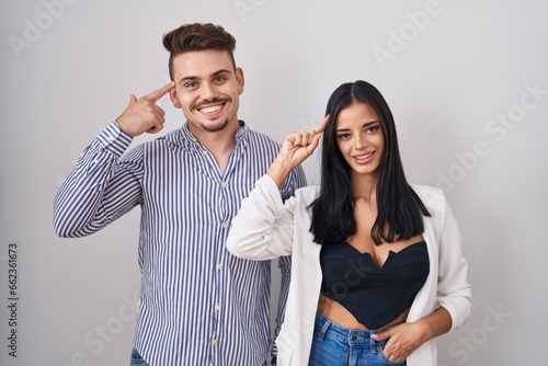 Young hispanic couple standing over white background smiling pointing to head with one finger, great idea or thought, good memory