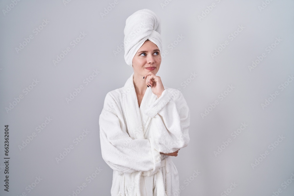 Blonde caucasian woman wearing bathrobe with hand on chin thinking about question, pensive expression. smiling and thoughtful face. doubt concept. Blonde caucasian woman wearing bathrobe with hand on chin thinking about question, pensive expression. smiling and thoughtful face. doubt concept.
