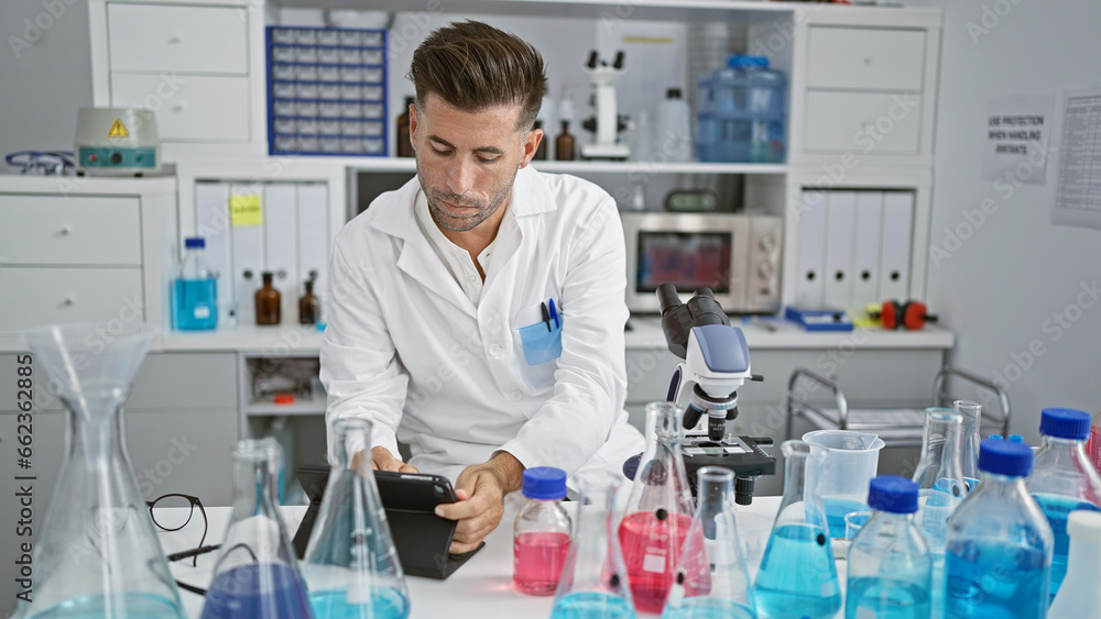 Fototapeta premium Handsome young hispanic man seriously concentrating as scientist, discovering through research on touchpad, sitting indoors at medical laboratory table.