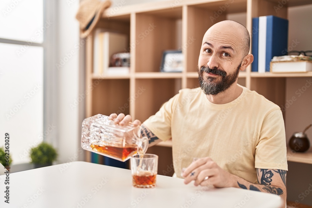 Young bald man pouring whisky on glass sitting on table at home Young bald man pouring whisky on glass sitting on table at home