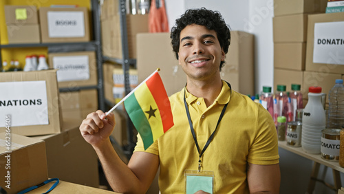 Young latin man volunteer holding ghana flag smiling at charity center