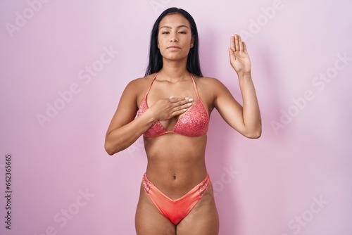 Hispanic woman wearing bikini swearing with hand on chest and open palm, making a loyalty promise oath