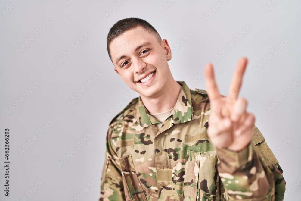 Young man wearing camouflage army uniform smiling looking to the camera showing fingers doing victory sign. number two. Young man wearing camouflage army uniform smiling looking to the camera showing fingers doing victory sign. number two.