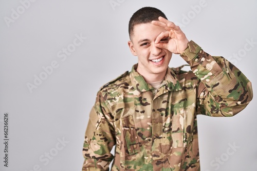 Young man wearing camouflage army uniform doing ok gesture with hand smiling  eye looking through fingers with happy face.