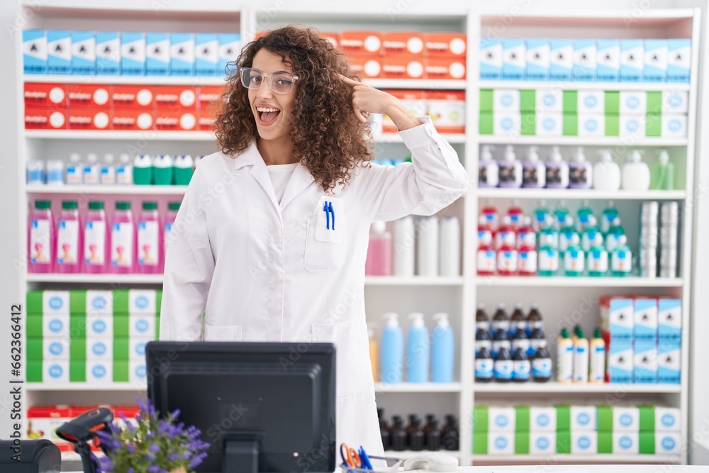 Hispanic woman with curly hair working at pharmacy drugstore smiling pointing to head with one finger, great idea or thought, good memory