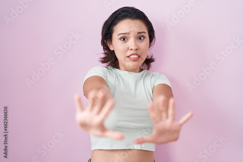 Hispanic young woman standing over pink background afraid and terrified with fear expression stop gesture with hands, shouting in shock. panic concept. © Krakenimages.com