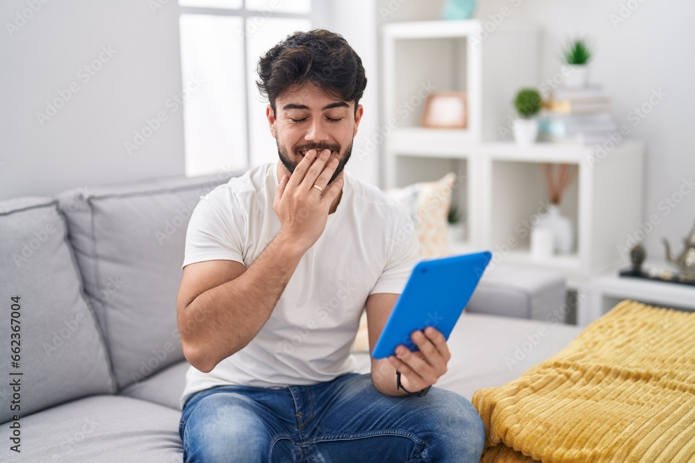 Hispanic man with beard using touchpad sitting on the sofa laughing and embarrassed giggle covering mouth with hands, gossip and scandal concept