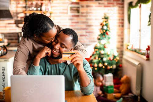 Young African American couple online shopping for presents on a laptop at their home decorated for the Christmas and new year holidays photo