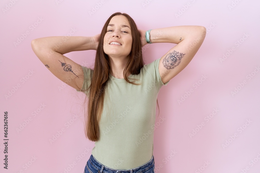 Beautiful brunette woman standing over pink background relaxing and stretching, arms and hands behind head and neck smiling happy Beautiful brunette woman standing over pink background relaxing and stretching, arms and hands behind head and neck smiling happy
