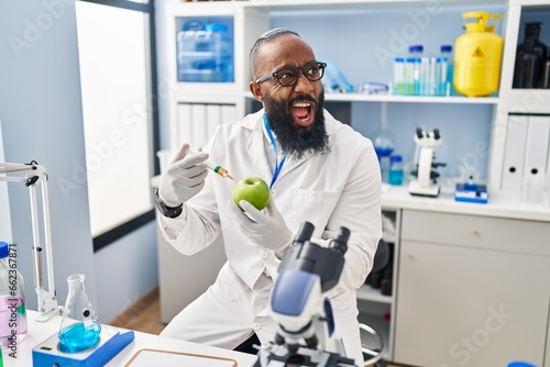 African american man working at scientist laboratory with apple angry and mad screaming frustrated and furious, shouting with anger. rage and aggressive concept.