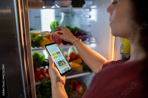 Mixed middle aged woman using her diet app on her smartphone while looking through her fridge at home photo