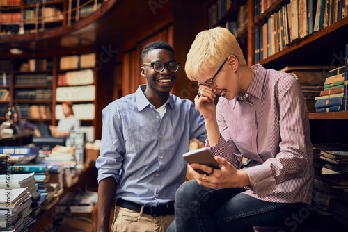 Diverse student duo laughing at a smartphone in a college library photo