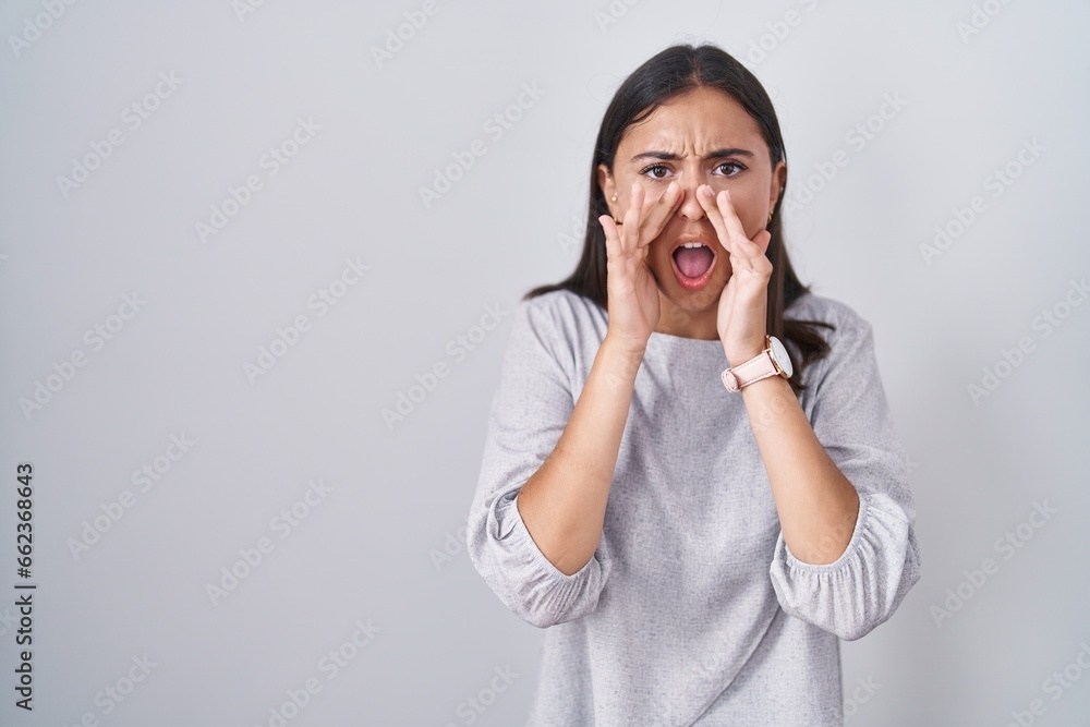 Young hispanic woman standing over white background shouting angry out loud with hands over mouth Young hispanic woman standing over white background shouting angry out loud with hands over mouth