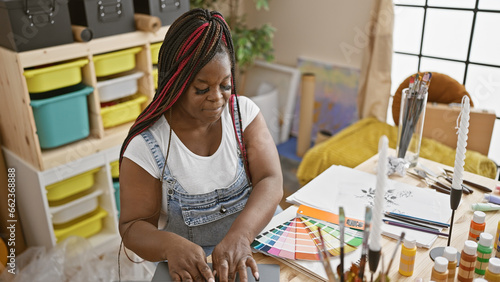 Inside the art studio, beautiful african american woman artist with braids confidently choosing color on laptop while immersed in her painting hobby