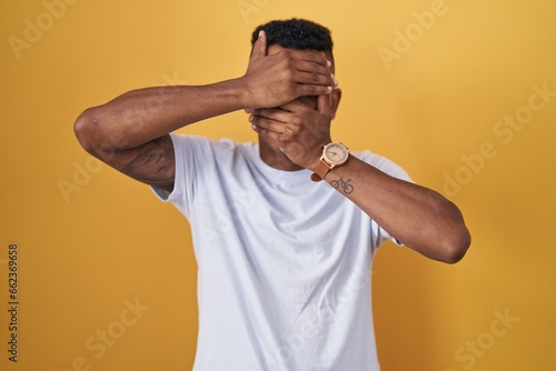 Young hispanic man standing over yellow background covering eyes and mouth with hands, surprised and shocked. hiding emotion
