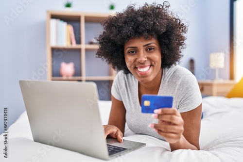 African american woman using laptop and credit card sitting on bed at bedroom