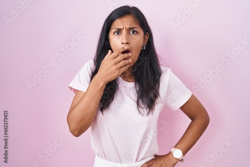 Young hispanic woman standing over pink background looking fascinated with disbelief, surprise and amazed expression with hands on chin