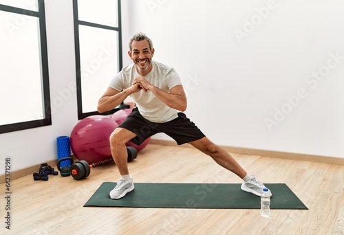 Middle age grey-haired man smiling confident stretching leg at sport center