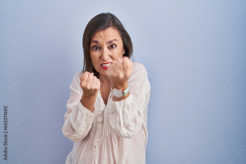 Middle age hispanic woman standing over blue background ready to fight with fist defense gesture, angry and upset face, afraid of problem Middle age hispanic woman standing over blue background ready to fight with fist defense gesture, angry and upset face, afraid of problem