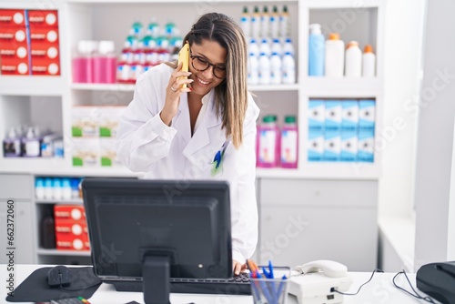 Young hispanic woman pharmacist talking on smartphone using computer at pharmacy