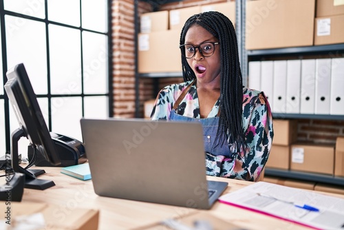 African woman with braids working at small business ecommerce with laptop scared and amazed with open mouth for surprise, disbelief face