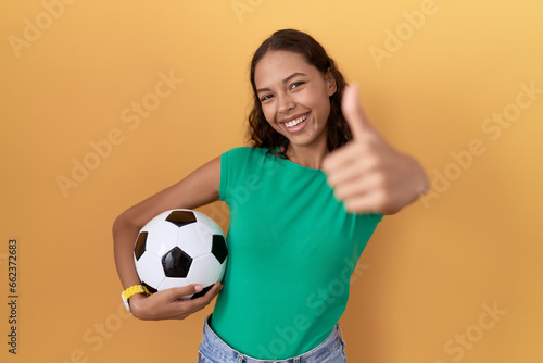 Young hispanic woman holding ball approving doing positive gesture with hand, thumbs up smiling and happy for success. winner gesture. © Krakenimages.com