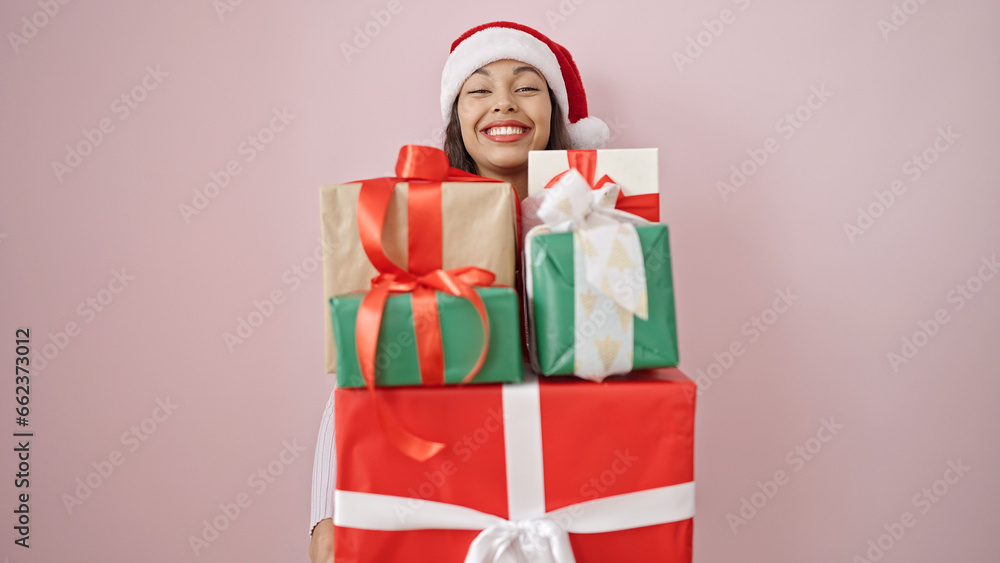 Young beautiful hispanic woman wearing christmas hat holding gifts over isolated pink background Young beautiful hispanic woman wearing christmas hat holding gifts over isolated pink background