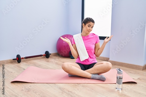 Young brunette woman sitting on yoga mat at the gym clueless and confused expression with arms and hands raised. doubt concept.
