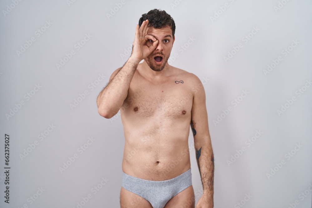 Fototapeta premium Young hispanic man standing shirtless wearing underware doing ok gesture shocked with surprised face, eye looking through fingers. unbelieving expression.