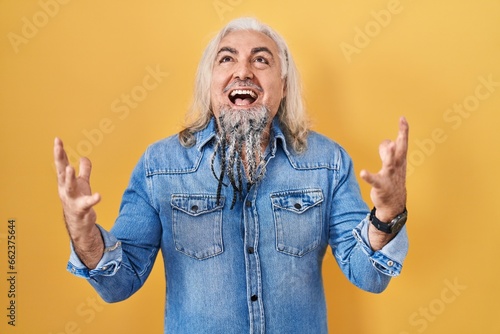 Middle age man with grey hair standing over yellow background celebrating mad and crazy for success with arms raised and closed eyes screaming excited. winner concept