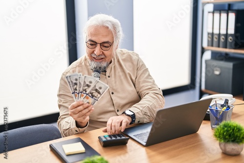 Middle age grey-haired man business worker using laptop counting dollars at office