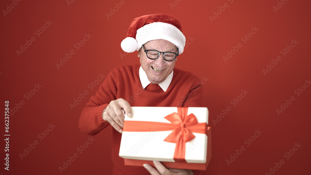 senior wearing christmas hat unpacking gift over isolated red background senior wearing christmas hat unpacking gift over isolated red background