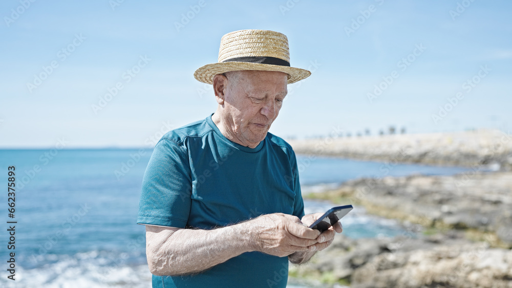 Senior grey-haired man tourist wearing summer hat using smartphone at seaside