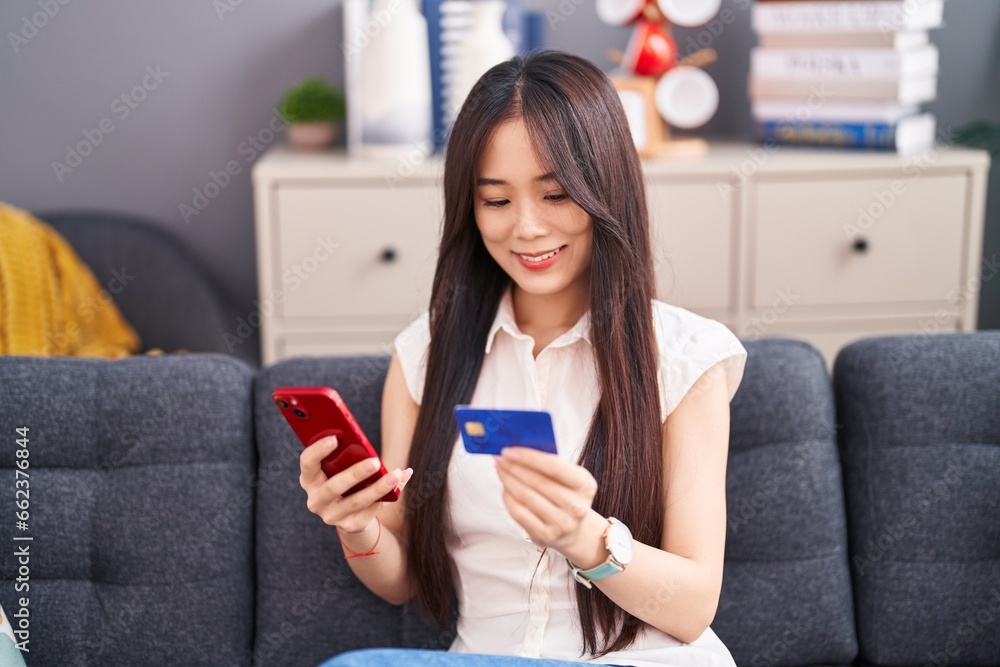 Young chinese woman using smartphone and credit card sitting on sofa at home