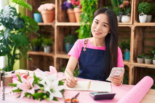 Young chinese woman florist counting dollars writing on notebook at flower shop