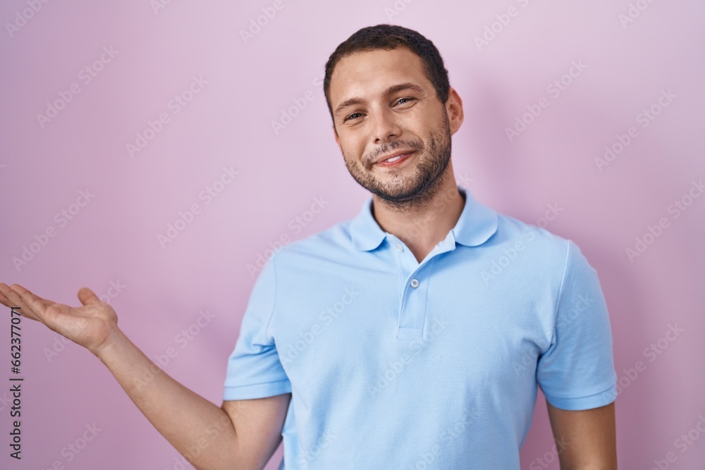 Fototapeta premium Hispanic man standing over pink background smiling cheerful presenting and pointing with palm of hand looking at the camera.